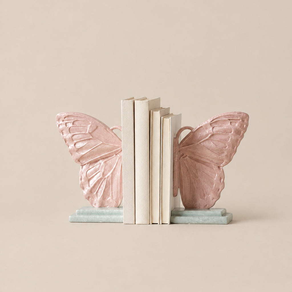 A pair of butterfly-shaped bookends in a soft blush pink color, holding a stack of neutral-colored books on a light gray background.