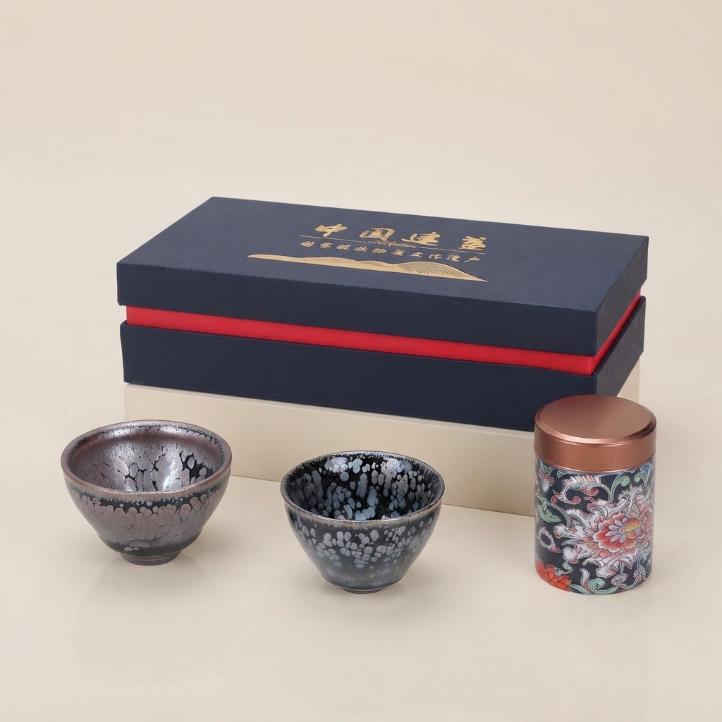 A set of ceramic tea ware and packaging on a warm cream background, featuring two small speckled tea bowls and a patterned cylindrical tin with a copper-toned lid, positioned in front of a dark navy presentation box with gold lettering and a red band.