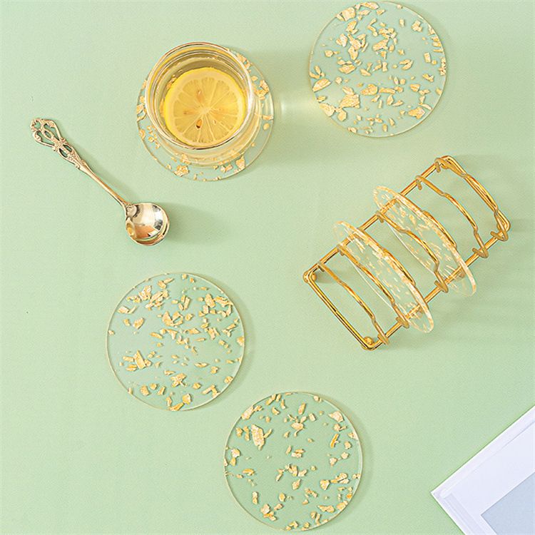 A set of clear round coasters with embedded gold leaf flakes, shown on a pale green background with a gold spoon, a glass of lemon tea, and a gold drink holder.