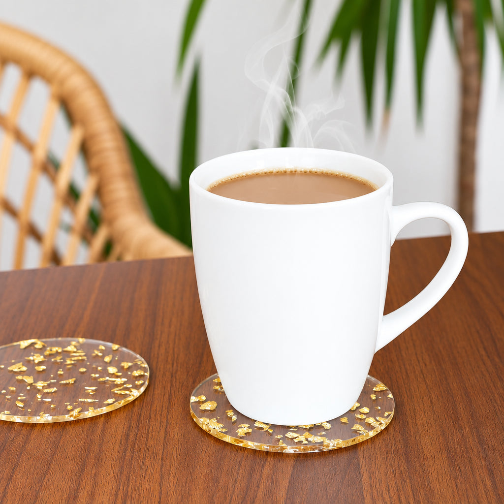 A white ceramic mug filled with a hot beverage sits on a round clear coaster with gold glitter flakes on a wooden table. A second matching coaster is placed beside it, and one coaster is shown alone in the foreground.