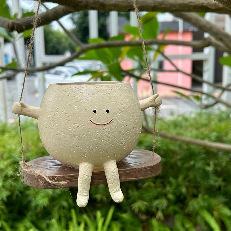A cute, whimsical ceramic hanging planter shaped like a smiling character with arms and legs, sitting on a wooden swing seat suspended by twine from a tree branch.